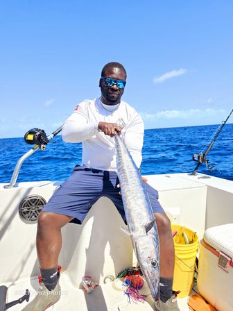 Offshore angler on a white boat holding a long silver wahoo (kingfish) with bright blue ocean and sky, sportfishing rods and gear visible.