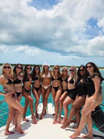Ten women in bikinis smiling and wearing sunglasses as they pose on the bow of a boat over turquoise tropical water beneath a dramatic cloudy blue sky