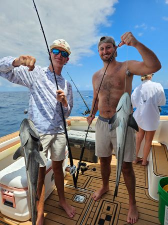 Two anglers on a fishing boat deck holding up large silver saltwater fish they just caught on an offshore deep-sea fishing trip under a blue sky and calm ocean.