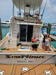 Smiling woman holding a bright green-yellow mahi-mahi on the teak deck of a sportfishing yacht docked at a Bahamas marina, with fishing rods and friends in the background.