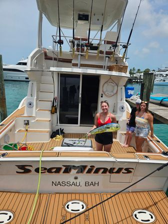 Smiling woman holding a bright green-yellow mahi-mahi on the teak deck of a sportfishing yacht docked at a Bahamas marina, with fishing rods and friends in the background.