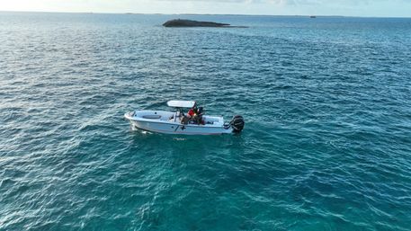 Small center-console fishing boat with anglers drifting on vibrant turquoise tropical ocean near a low rocky islet under a clear sky