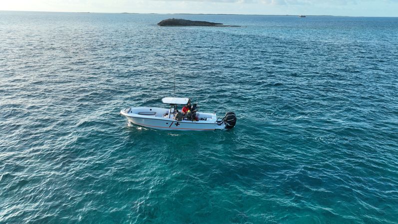 Small center-console fishing boat with anglers drifting on vibrant turquoise tropical ocean near a low rocky islet under a clear sky