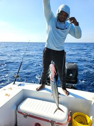 Angler on a small boat in bright offshore blue water holding a freshly caught mackerel on a colorful lure above a white cooler — sunny deep-sea fishing scene.
