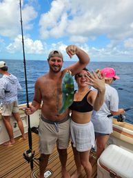 Smiling anglers on an offshore fishing boat holding a freshly caught vibrant green-blue parrotfish against a sunny, partly cloudy tropical sea backdrop