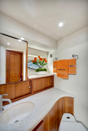 Cozy yacht bathroom head with curved white countertop and oval sink, warm wood cabinetry, round porthole window, orange tulips in a white planter, matching orange towels on a chrome rack and a compact marine toilet.