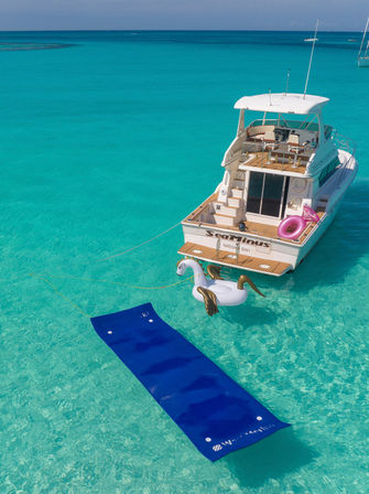 White motor yacht anchored in crystal-clear turquoise tropical water with a long blue floating mat, a gold-tipped inflatable swan and a pink swim ring tied to the stern.