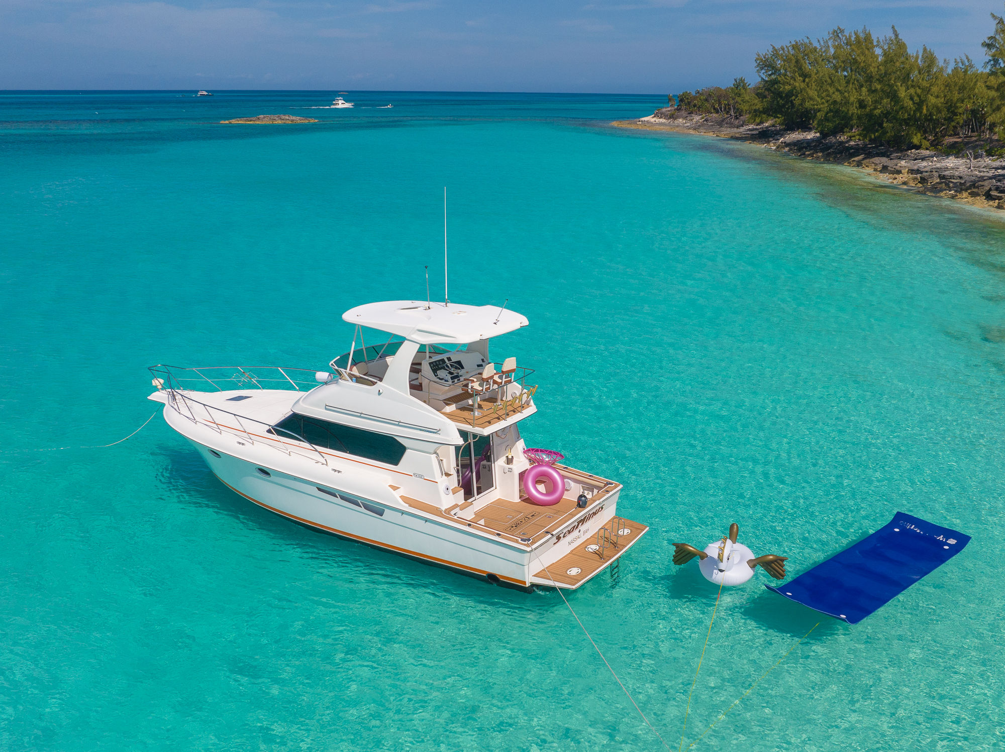 White motor yacht anchored in shallow turquoise tropical lagoon near a small tree-lined islet, surrounded by a pink donut float, a unicorn float and a blue water lounger.