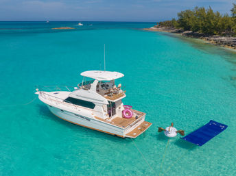 White motor yacht anchored in shallow turquoise tropical lagoon near a small tree-lined islet, surrounded by a pink donut float, a unicorn float and a blue water lounger.