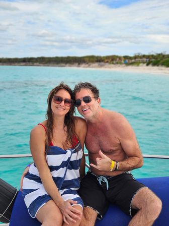 Smiling couple on a boat over turquoise water near a sandy tropical coastline; woman in sunglasses wrapped in a blue-and-white striped towel and man shirtless in sunglasses and swim trunks flashing a shaka sign on a sunny vacation.