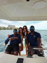 Four people enjoying a motorboat cruise near a coastal skyline — two women in bikinis smiling between two men in navy shirts steering the boat, blue ocean and shoreline buildings in the background.