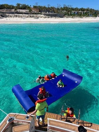 Aerial view of kids and adults in bright life jackets playing on a blue inflatable water mat beside a boat in clear turquoise tropical waters off a sandy, palm‑lined beach.