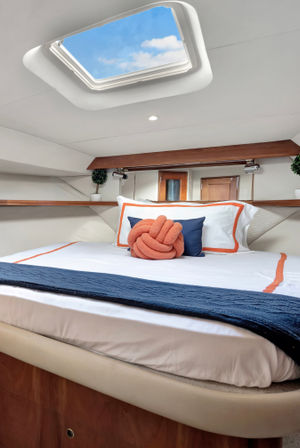 Cozy yacht cabin bedroom with open skylight showing blue sky, white bed dressed in navy and orange-trim linens, navy throw and orange knot pillow, wood trim and small potted plants