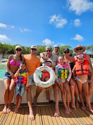 Smiling multi-generation family in colorful swimsuits and life jackets on a boat deck, holding a life ring labeled Nassau, BHS, with turquoise Bahamian water and a rocky shoreline under a bright blue sky.