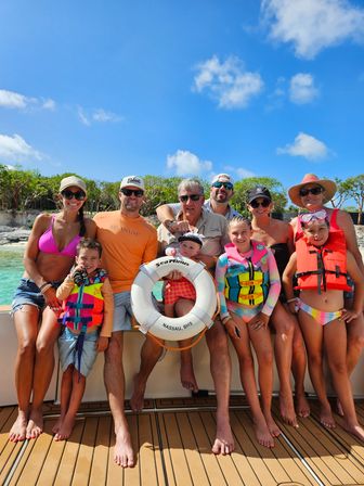 Smiling multi-generation family in colorful swimsuits and life jackets on a boat deck, holding a life ring labeled Nassau, BHS, with turquoise Bahamian water and a rocky shoreline under a bright blue sky.