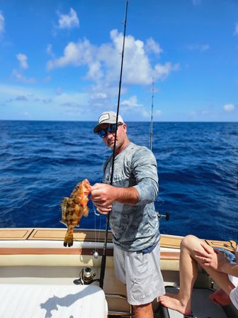 Angler wearing a cap and sunglasses holding a freshly caught brown-spotted grouper on a boat over deep-blue offshore waters with fishing rods and sunny sky in the background.