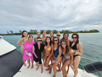 Smiling group of women in swimsuits posing on a white boat deck with turquoise ocean water and a tree-lined tropical island under a cloudy sky — lively coastal boat day trip.