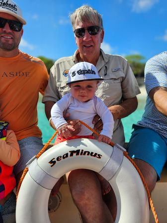 Smiling baby in a white cap and sun shirt sits on a man’s lap on a small boat, holding a white lifebuoy with orange rope against turquoise water and a bright blue sky.