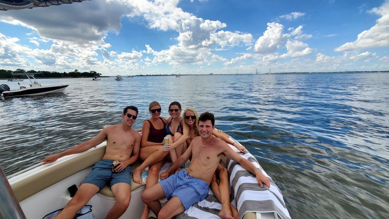 Five young adults relaxing on the bow of a motorboat in a sunny coastal bay, holding drinks with calm water, blue sky, scattered clouds and a distant bridge on the horizon.
