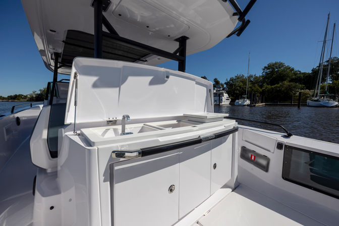White boat cockpit wet bar with sink, faucet, counter and storage compartments under an open hatch, docked at a marina with sailboats and trees under a clear blue sky.
