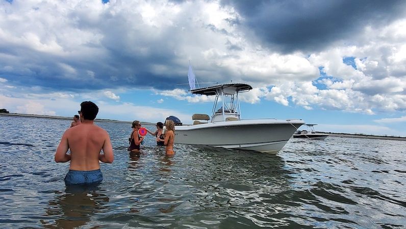 Group of people wading in shallow coastal water beside a white center-console boat under a dramatic cloudy blue sky — summer boating near shore.