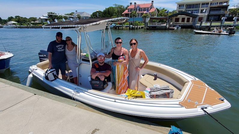 Five adults enjoying a sunny outing on a white center-console boat moored at a marina, with beach bags and towels on deck and coastal homes lining the calm water in the background.