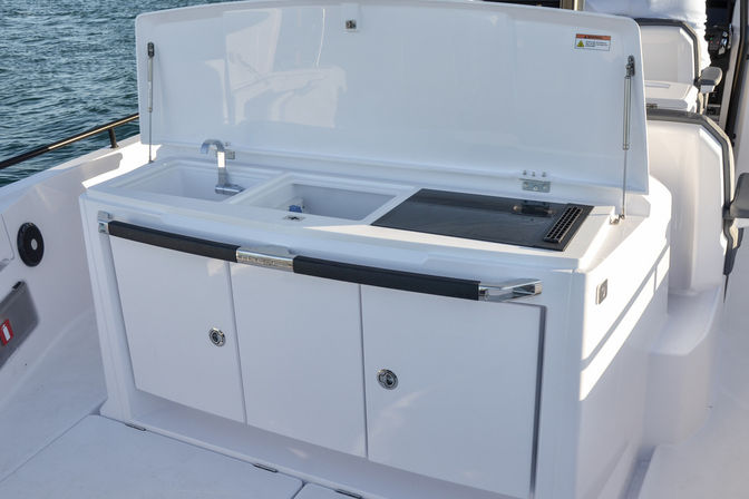White boat galley on deck with open hatch revealing twin sinks and built-in cooktop, ocean water in the background