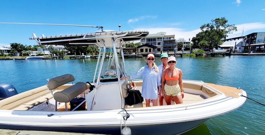 Three people in summer outfits standing on a center-console motorboat with T-top, docked at a sunny coastal marina with waterfront homes and calm blue water