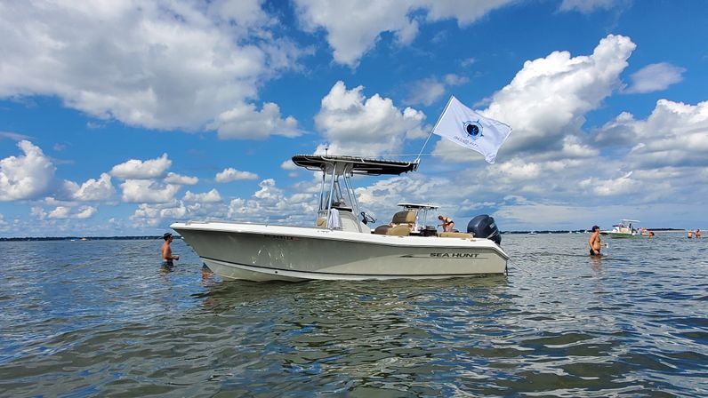 White center-console boat anchored in shallow coastal bay on a sunny summer day, people wading in calm water under a blue sky with puffy clouds and a fluttering white flag
