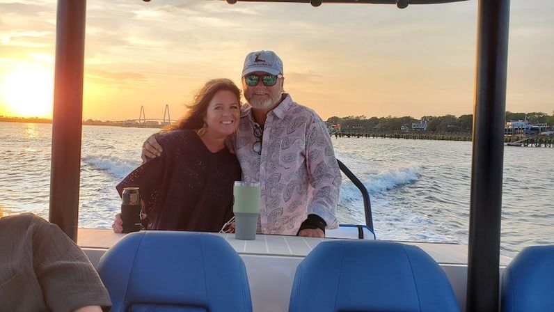 Couple on a sunset cruise hugging on a boat with drinks, blue seats in the foreground, wake trailing across coastal waters past a marina and bridge.
