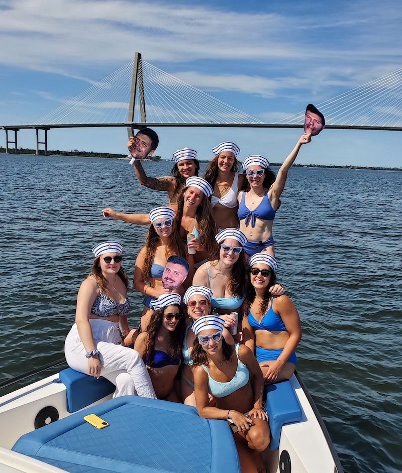 Group boat party of women in sailor hats and bikinis smiling on a sunny day on the water with a cable-stayed bridge spanning the bay in the background