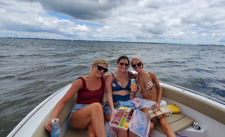 Three friends in swimsuits and sunglasses on a small boat, smiling and holding drinks with a charcuterie-style snack box, choppy open water and cloudy sky behind them — summer boating outing.