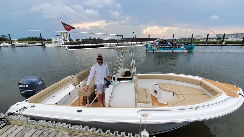 Person in a sun hat aboard a white center-console boat with tan seating docked at a coastal marina under a moody sunset sky, other boats passing in the background.