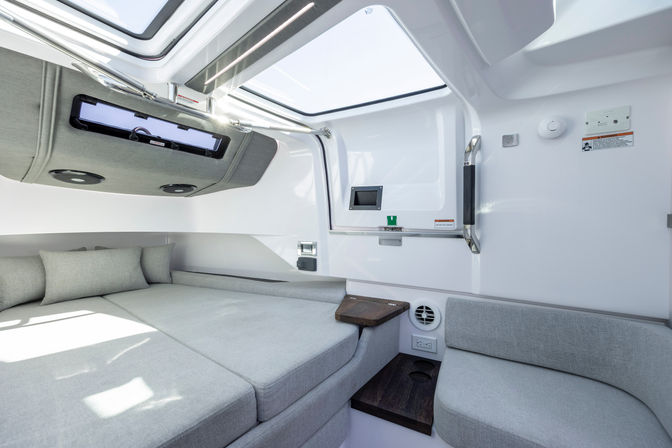 Cozy bright yacht cabin interior with light-gray V-berth bed and cushions, overhead skylight hatch, built-in seating, small wooden side tables, stainless handrail and modern marine fittings