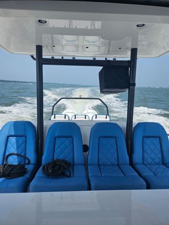Aft view from a powerboat with four bright blue cushioned seats, coiled ropes, T-top overhead and a foamy wake stretching across open coastal water under a clear sky.