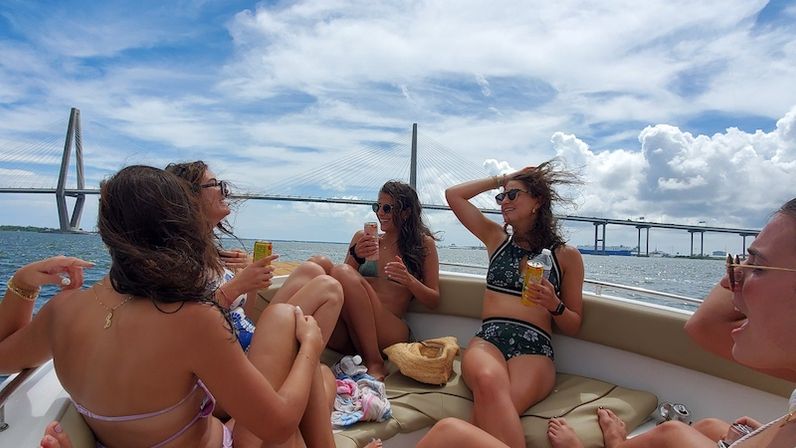 Friends in bikinis laughing with cold drinks on a sunlit motorboat cruising past a large cable-stayed bridge over blue water and a coastal skyline