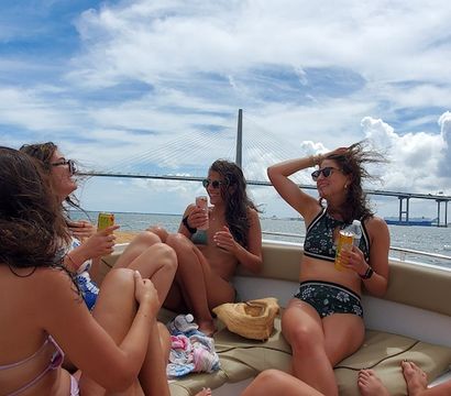 Friends in bikinis laughing with cold drinks on a sunlit motorboat cruising past a large cable-stayed bridge over blue water and a coastal skyline