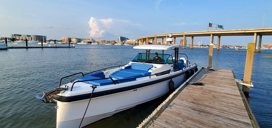White motorboat with blue cushions docked at a wooden marina pier on calm harbor water, sunny waterfront with buildings and an arched highway bridge in the background