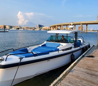 White motorboat with blue cushions docked at a wooden marina pier on calm harbor water, sunny waterfront with buildings and an arched highway bridge in the background