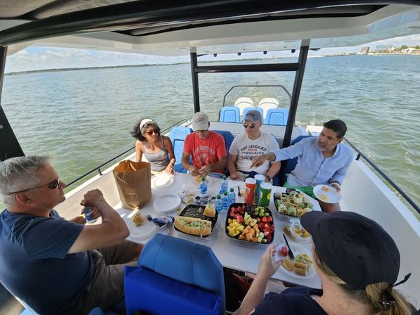 Group of adults enjoying a casual boat picnic aboard a motorboat on a sunny coastal bay, with sandwiches, fruit platter and drinks on the table.