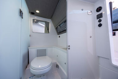 Compact white marine bathroom (boat head) with round toilet, countertop storage vent, angled mirror, overhead LED light and nearby electrical control panel — sleek yacht interior.