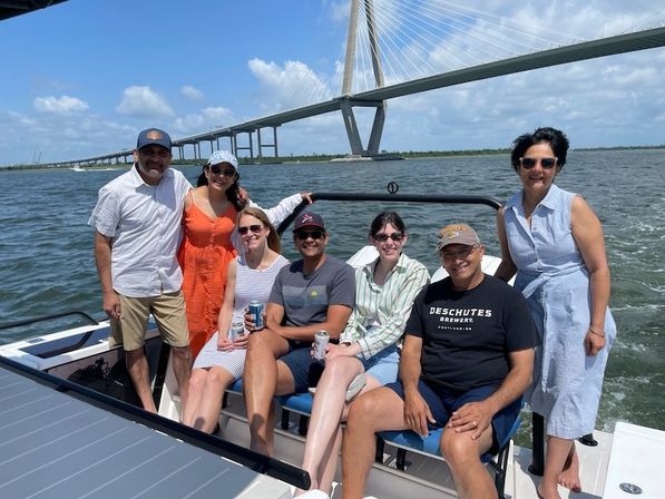 Seven friends smiling and relaxing on a motorboat under a large cable-stayed bridge on a sunny summer day, blue water and sky with scattered clouds in the background.