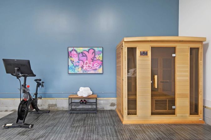 Indoor fitness room with a stationary exercise bike, wooden sauna cabin, bench holding rolled towels and dumbbells, and colorful “Love” wall art on a blue accent wall — cozy wellness space.
