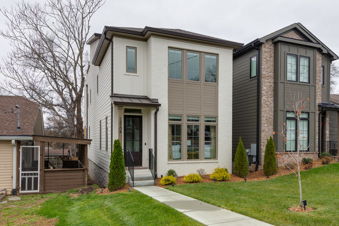 Modern two-story suburban townhouse with white brick façade and large grid windows, small covered entry and concrete walkway, manicured lawn, leafless trees and a dark gray neighboring home on an overcast day.