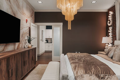Modern urban bedroom with a gold fringe chandelier, velvet brown bedding, dark accent wall, wooden media console with wall TV, and frosted sliding door to a kitchenette.