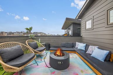 Cozy rooftop patio with a modern L-shaped outdoor sectional, two wicker lounge chairs, a round fire pit on a colorful pastel rug, potted plant and distant urban skyline.