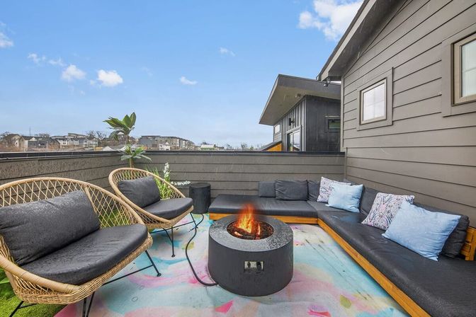 Cozy rooftop patio with a modern L-shaped outdoor sectional, two wicker lounge chairs, a round fire pit on a colorful pastel rug, potted plant and distant urban skyline.