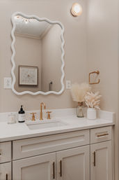 Modern neutral powder-room vanity with wavy white mirror, brass faucet and hardware, white quartz countertop, pampas grass in vases and minimalist soap and cotton accessories.