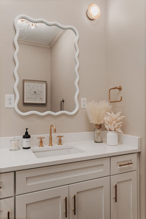 Modern neutral powder-room vanity with wavy white mirror, brass faucet and hardware, white quartz countertop, pampas grass in vases and minimalist soap and cotton accessories.