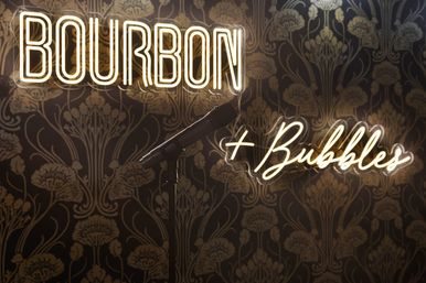 Neon-lit 'BOURBON + Bubbles' sign on patterned wallpaper with a microphone on a stand, moody bar stage setting for live music and cocktails.